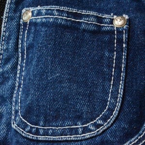 May include: Close-up of a denim pocket with white stitching and two silver buttons. The pocket is a deep indigo blue, with a curved bottom and a rectangular shape. The buttons have a decorative design.