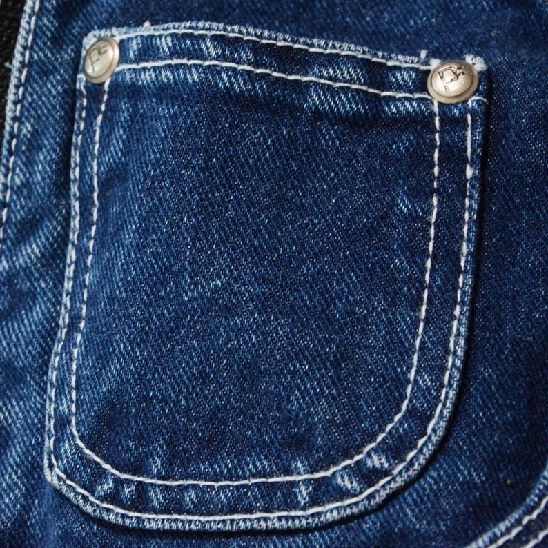 May include: Close-up of a denim pocket with white stitching and two silver buttons. The pocket is a deep indigo blue, with a curved bottom and a rectangular shape. The buttons have a decorative design.
