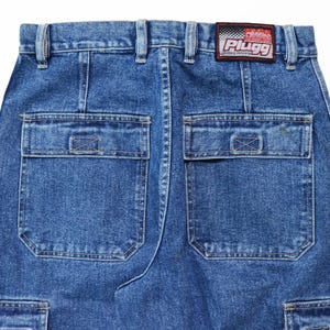 May include: Close-up of the back of blue denim cargo pants. The pants have two large square pockets on the back, with a smaller pocket on the side. A red and white patch with the text "Plugg Clothing Company" is sewn on the waistband.