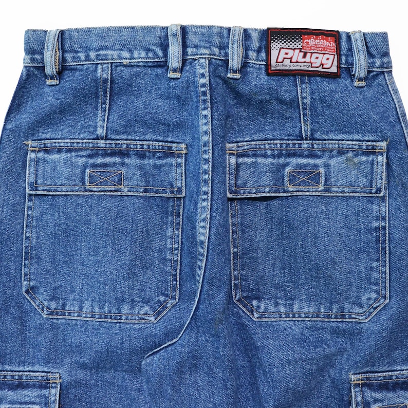 May include: Close-up of the back of blue denim cargo pants. The pants have two large square pockets on the back, with a smaller pocket on the side. A red and white patch with the text "Plugg Clothing Company" is sewn on the waistband.