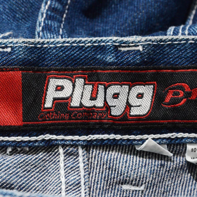 May include: Close-up of a blue denim garment with white stitching. A black and red label displays the brand name "Plugg" in white lettering. The label also includes the text "Clothing Company."