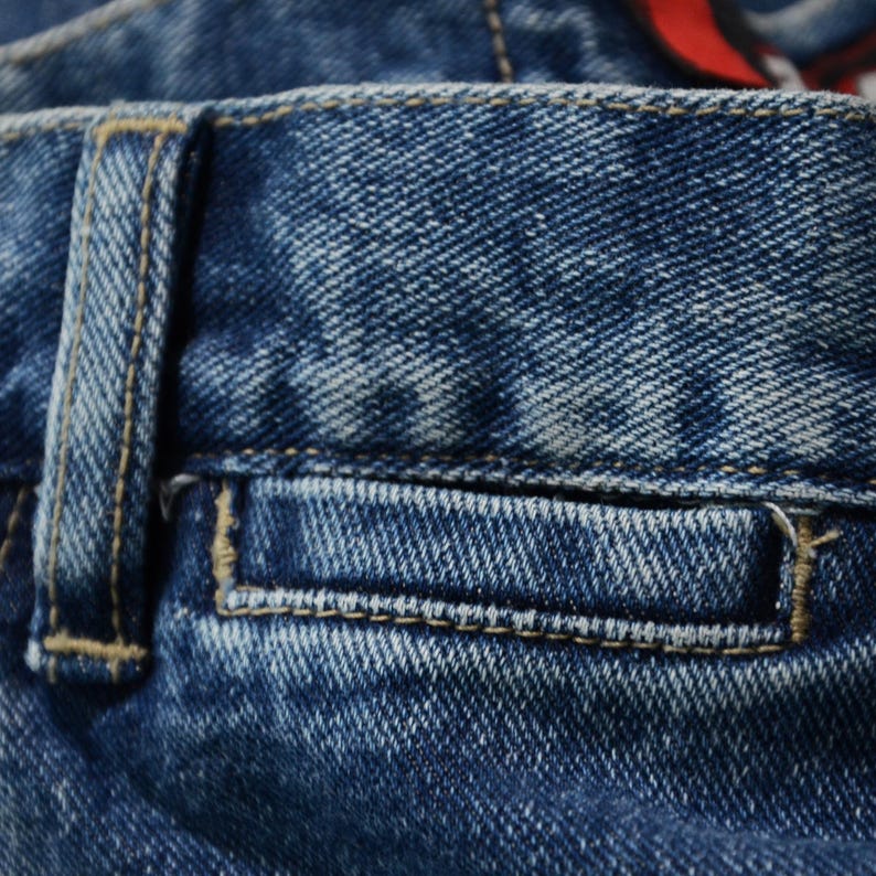 May include: Close-up of blue denim jeans, showcasing the pocket and belt loop details. The denim has a faded, worn appearance with visible stitching in a contrasting tan color. The texture of the fabric is clearly visible.