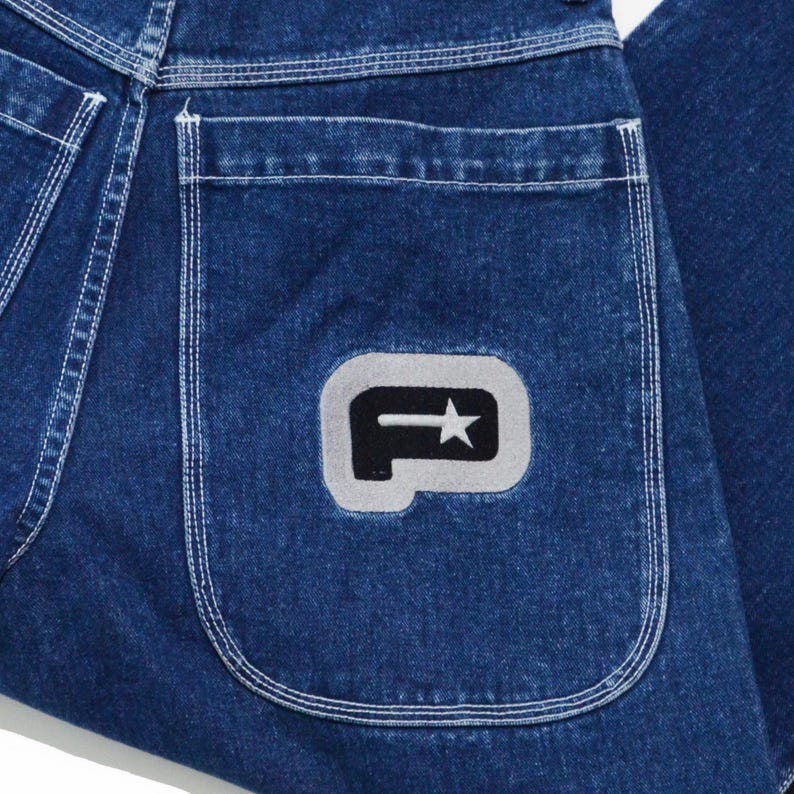 May include: Close-up of blue denim jeans with a white stitched pocket. The pocket features a gray and white logo with a star. The jeans have a classic five-pocket design.