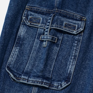 May include: Close-up of a section of blue denim fabric featuring a large pocket with a flap and a smaller pocket above it. The pockets have decorative stitching and a buckle closure. The denim has a slightly faded appearance.