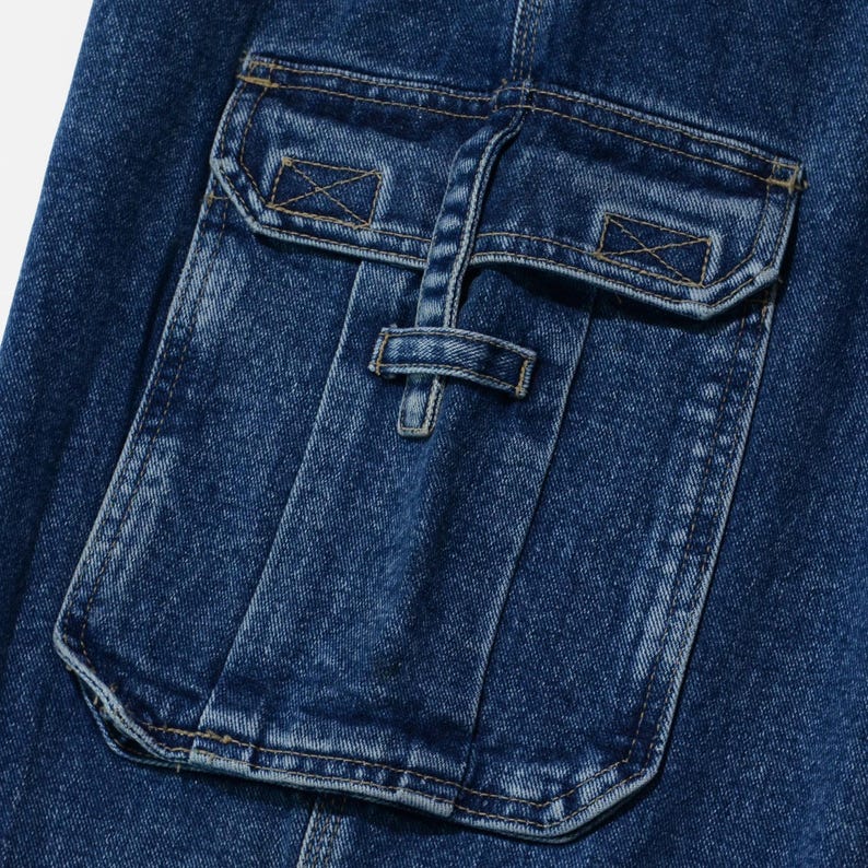 May include: Close-up of a section of blue denim fabric featuring a large pocket with a flap and a smaller pocket above it. The pockets have decorative stitching and a buckle closure. The denim has a slightly faded appearance.