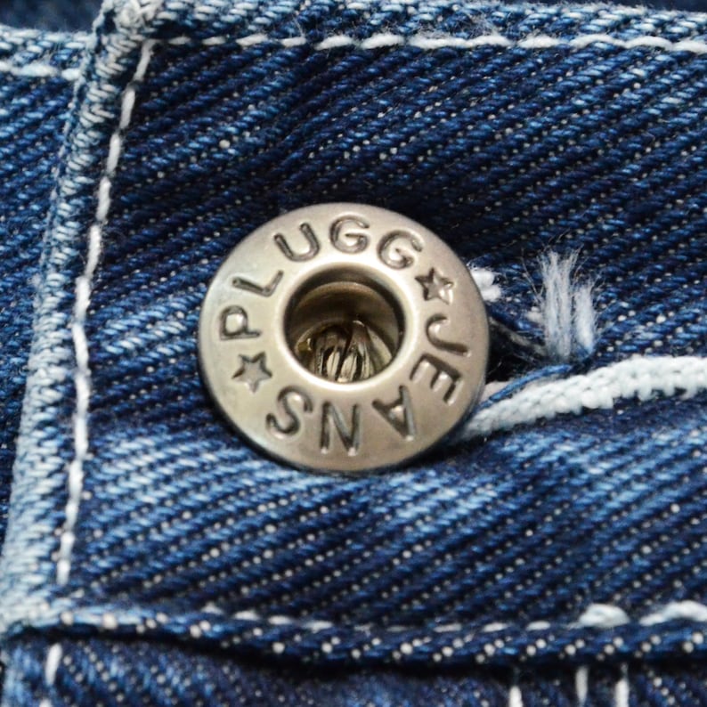 May include: Close-up of a silver button on blue denim jeans. The button is embossed with the words "PLUGG JEANS" around the edge and has a star design. White stitching is visible on the denim.