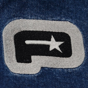 May include: Close-up of a denim fabric with an embroidered patch. The patch features a black shape with a white star and a horizontal line, outlined in gray. The background is a textured blue denim.