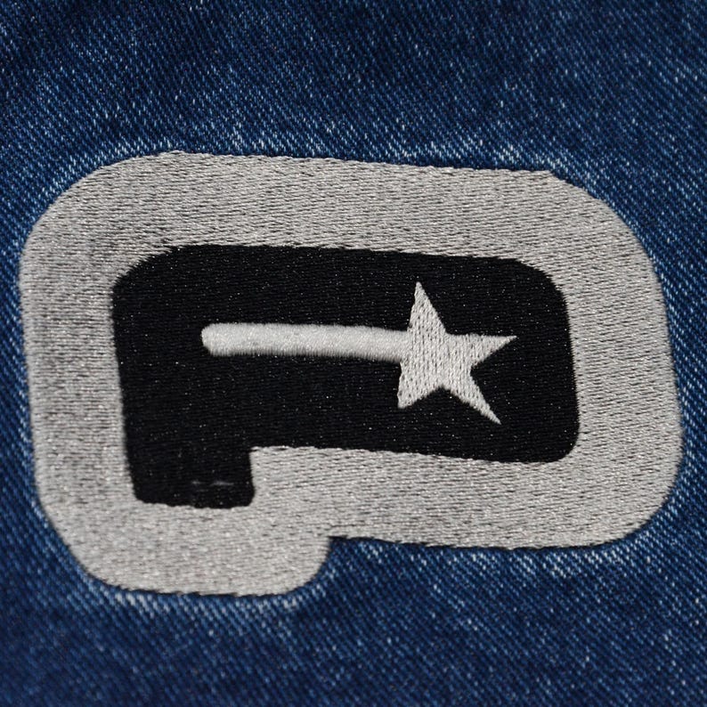 May include: Close-up of a denim fabric with an embroidered patch. The patch features a black shape with a white star and a horizontal line, outlined in gray. The background is a textured blue denim.