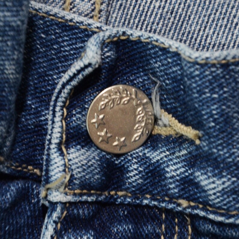 May include: Close-up of a silver button on blue denim jeans. The button has a circular design with three stars and a laurel wreath. The denim fabric shows visible stitching and a faded wash.