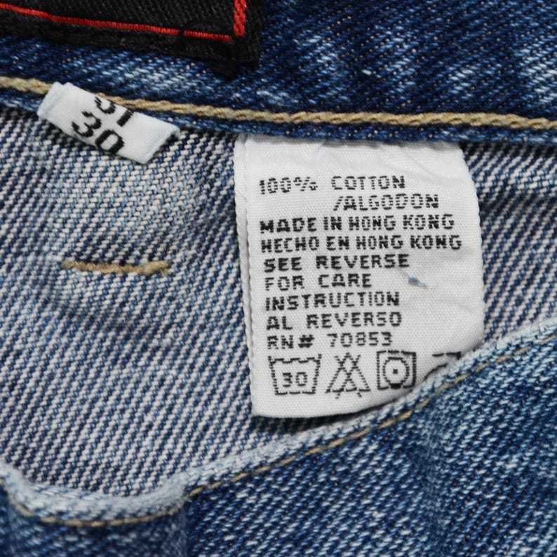 May include: Close-up of blue denim jeans with a white tag. The tag reads "100% COTTON / ALGODON MADE IN HONG KONG" and includes care instructions. A smaller tag shows the size "30".