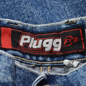 May include: Close-up of blue denim jeans. The jeans feature a black and red label with the word "Plugg" in white, and the text "Clothing Company" below. A small white tag displays the size "30".