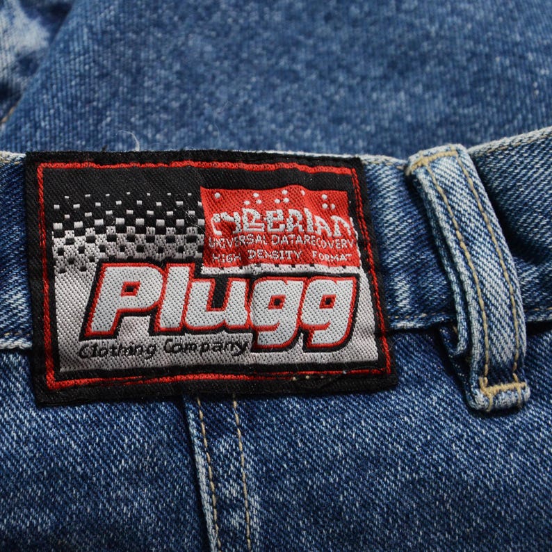 May include: Close-up of blue denim jeans with a rectangular label. The label is black with white text that reads "CYBERIAN UNIVERSAL DATARECOVERY HIGH DENSITY FORMAT". Below is the word "Plugg" in a white, outlined font. The words "Clothing Company" are at the bottom.