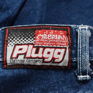 May include: Close-up of a blue denim jeans label. The label is rectangular with a black and white checkered pattern on the left side and the word "Plugg" in large red letters. The label also includes the text "Cyberian Universal Data Recovery High Density Format".