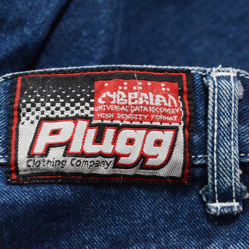 May include: Close-up of a blue denim jeans label. The label is rectangular with a black and white checkered pattern on the left side and the word "Plugg" in large red letters. The label also includes the text "Cyberian Universal Data Recovery High Density Format".