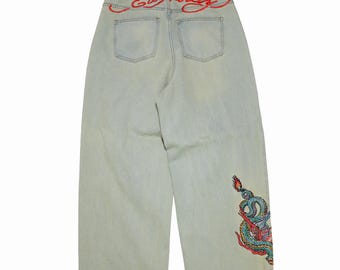 Może przedstawiać: Jasne dżinsy z szerokimi nogawkami. Z tyłu czerwony haft z napisem "Ed Hardy" i dwie kieszenie. Kolorowy wzór smoka jest wyhaftowany na prawej nogawce.
