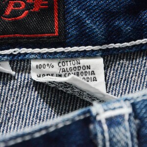 May include: Close-up of blue denim fabric with white stitching. A white tag reads "100% COTTON / ALGODON MADE IN CAMBODIA". A black tag with red lettering is visible in the upper left corner.
