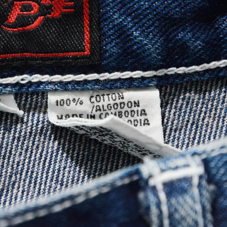 May include: Close-up of blue denim fabric with white stitching. A white tag reads "100% COTTON / ALGODON MADE IN CAMBODIA". A black tag with red lettering is visible in the upper left corner.