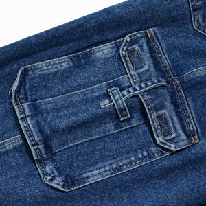 May include: Close-up of blue denim fabric with a large, rectangular pocket. The pocket features a flap secured by a strap and a button closure. The denim has a textured appearance with visible stitching details.