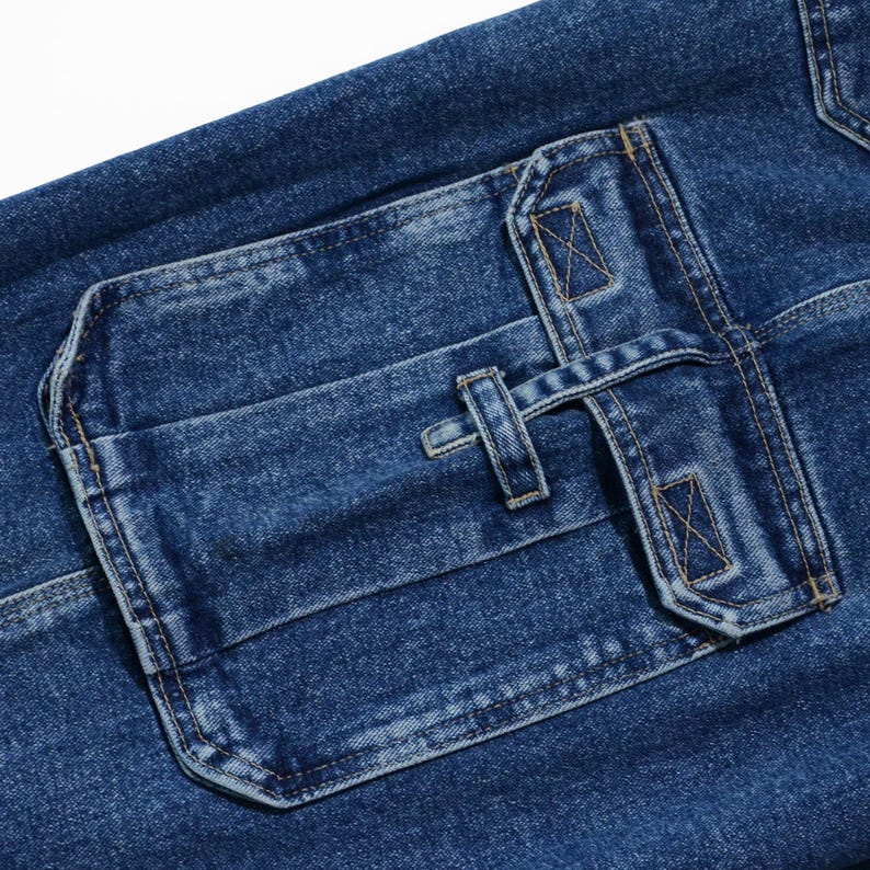May include: Close-up of blue denim fabric with a large, rectangular pocket. The pocket features a flap secured by a strap and a button closure. The denim has a textured appearance with visible stitching details.