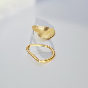 May include: Two gold-coloured earrings. One is a freeform shape, and the other is an open oval. The earrings are on a white surface. The earrings are made of a shiny metal.