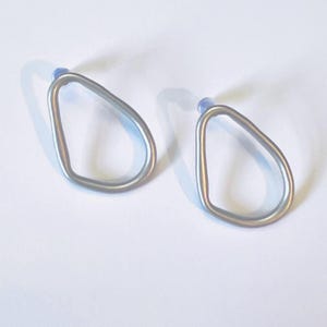 May include: Two silver-coloured, open-frame earrings. Each earring has a teardrop shape with a small, light blue post. The earrings are displayed on a white surface, casting a shadow.