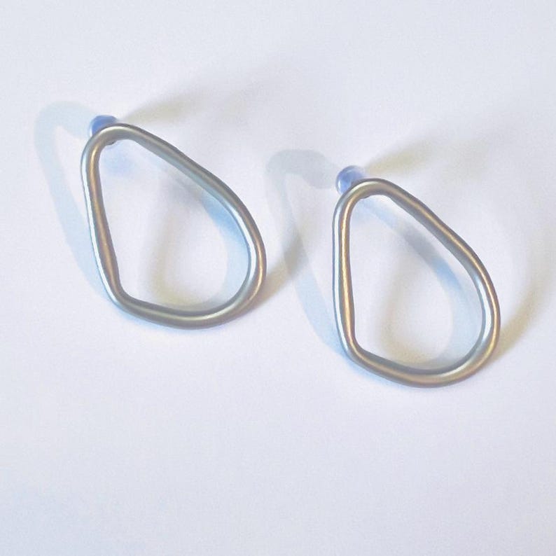 May include: Two silver-coloured, open-frame earrings. Each earring has a teardrop shape with a small, light blue post. The earrings are displayed on a white surface, casting a shadow.