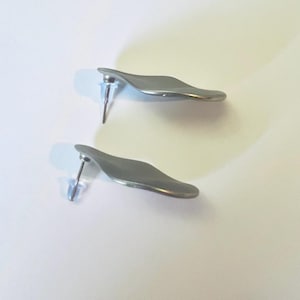 May include: A pair of silver-coloured stud earrings. Each earring features a curved, leaf-like design with a post and a clear plastic backing. The earrings are presented on a white surface.