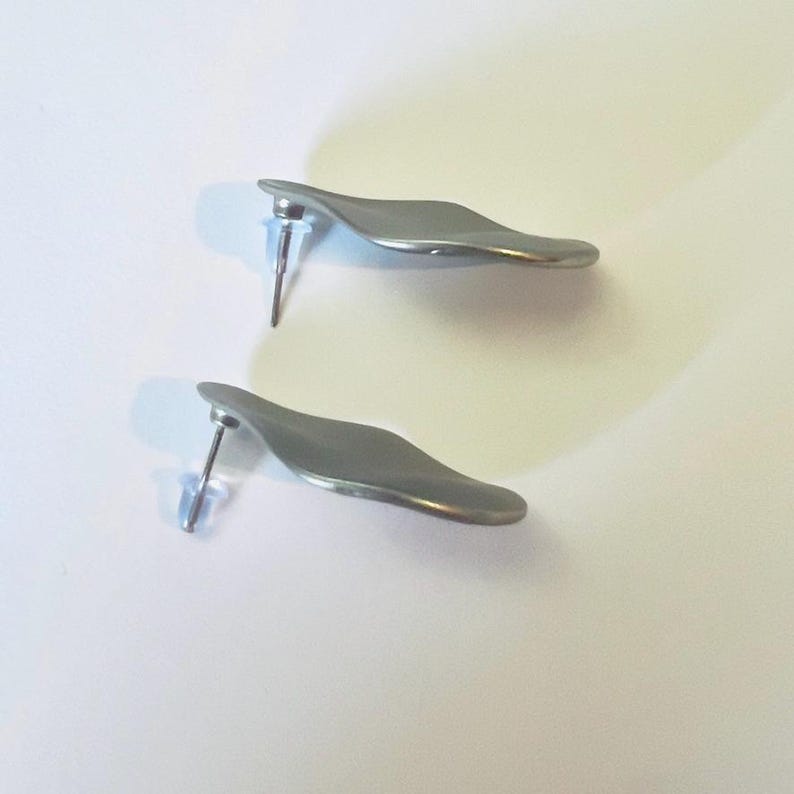 May include: A pair of silver-coloured stud earrings. Each earring features a curved, leaf-like design with a post and a clear plastic backing. The earrings are presented on a white surface.