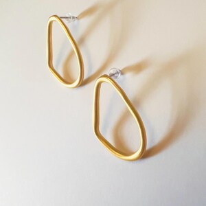 May include: A pair of gold-coloured, open-frame earrings. Each earring has a teardrop shape with a post for pierced ears. The earrings are displayed on a white surface, casting a soft shadow.