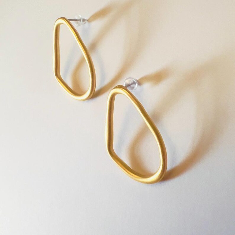 May include: A pair of gold-coloured, open-frame earrings. Each earring has a teardrop shape with a post for pierced ears. The earrings are displayed on a white surface, casting a soft shadow.