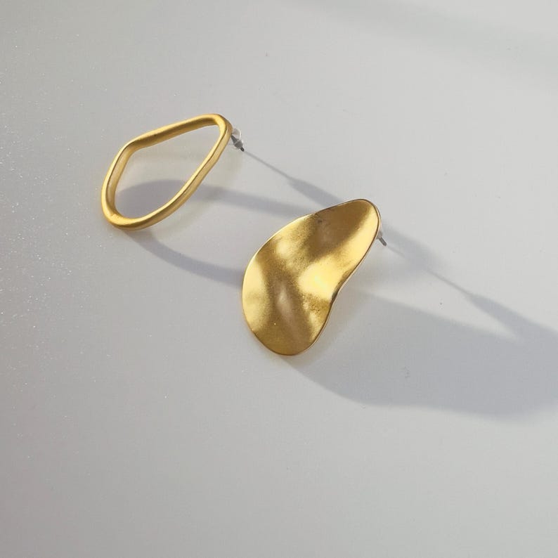 May include: A pair of gold-coloured earrings. One earring is an open, irregular oval shape. The other earring is a gold, organic, teardrop shape. Both earrings have a post for pierced ears. The earrings are on a light grey surface.