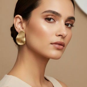 May include: Gold-coloured, abstract-shaped earrings. The earrings have a matte finish and a fluid, organic design. The image shows a close-up of the earrings on a person's ear, highlighting their shape and texture. The earrings are a statement piece.