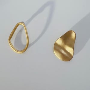 May include: A pair of gold earrings. One is an open, irregular shape, the other a solid, organic form. The earrings are displayed on a white surface, with a shadow cast behind them.