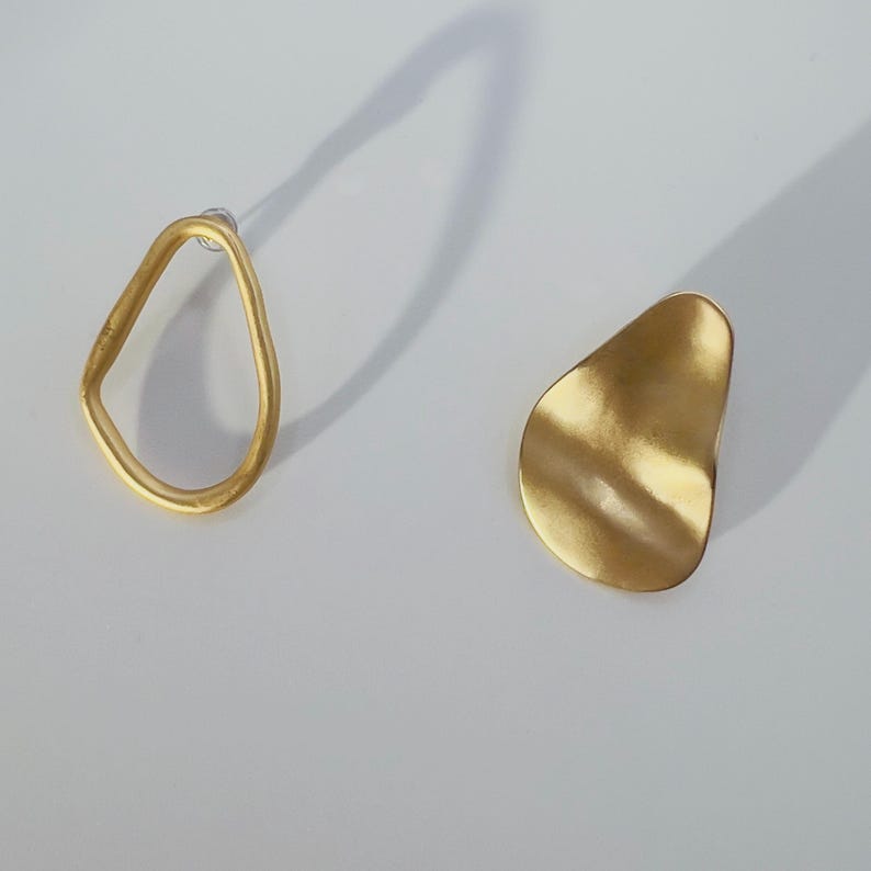 May include: A pair of gold earrings. One is an open, irregular shape, the other a solid, organic form. The earrings are displayed on a white surface, with a shadow cast behind them.