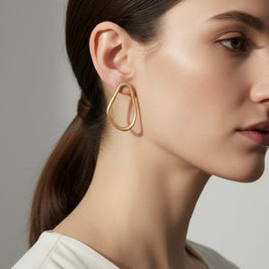 May include: Gold-coloured, abstract-shaped earrings. The earrings have a smooth, matte finish and a unique, irregular design. The earrings are worn on a person's ear, and the background is a neutral colour.