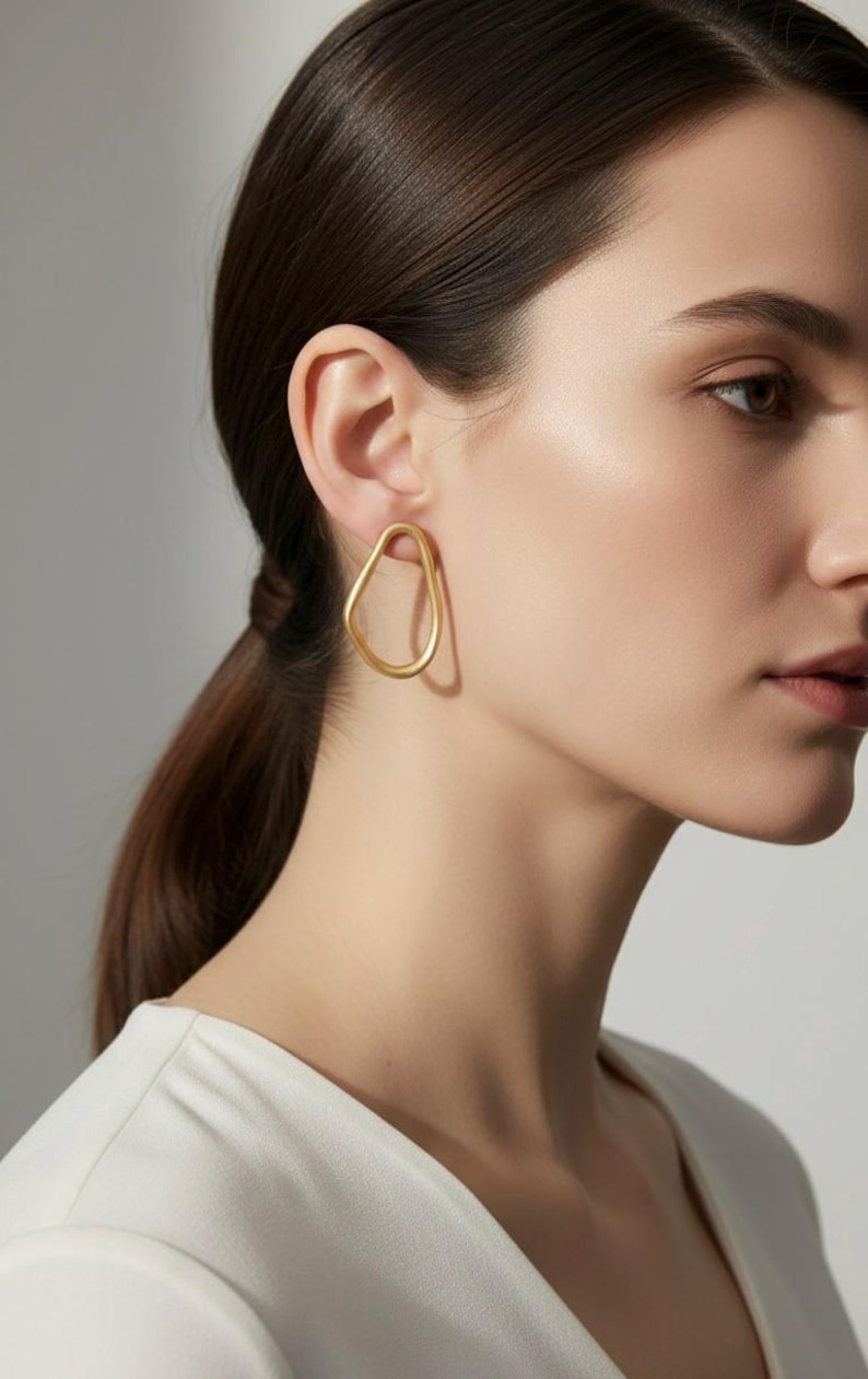 May include: Gold-coloured, abstract-shaped earrings. The earrings have a smooth, matte finish and a unique, irregular design. The earrings are worn on a person's ear, and the background is a neutral colour.