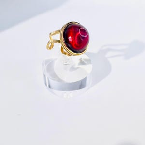 May include: A gold-toned adjustable ring featuring a large, round, red gemstone. The gemstone has a swirling, marbled appearance. The ring is displayed on a clear, cylindrical stand against a white background.