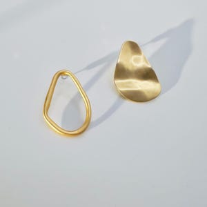 May include: A pair of gold-coloured earrings. One earring is a freeform, open shape, while the other is a solid, teardrop-shaped design with a textured surface. The earrings are displayed on a white background, casting a shadow.
