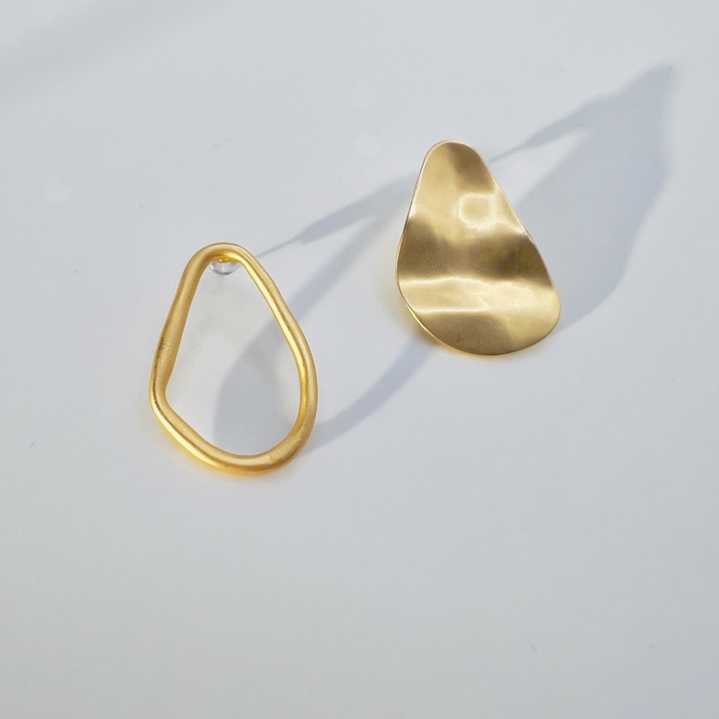 May include: A pair of gold-coloured earrings. One earring is a freeform, open shape, while the other is a solid, teardrop-shaped design with a textured surface. The earrings are displayed on a white background, casting a shadow.