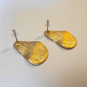 May include: A pair of gold-coloured stud earrings. Each earring has an irregular, leaf-like shape with a textured surface. The earrings have a post and backing for secure wear. The background is a light, neutral colour.