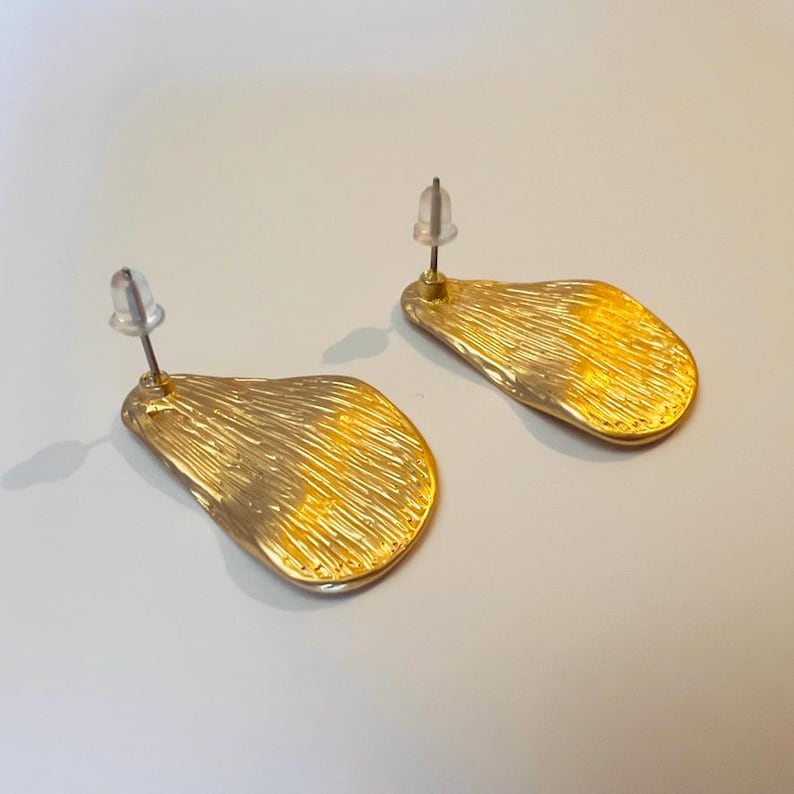 May include: A pair of gold-coloured stud earrings. Each earring has an irregular, leaf-like shape with a textured surface. The earrings have a post and backing for secure wear. The background is a light, neutral colour.
