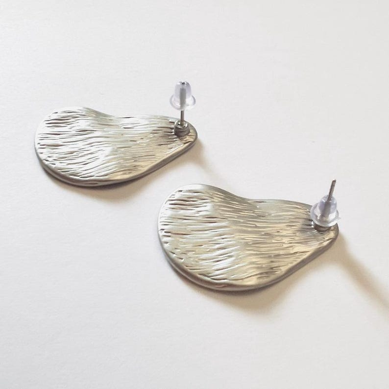 May include: A pair of silver stud earrings. Each earring features a textured, organic shape with a post and clear earring back. The earrings are presented on a white surface.