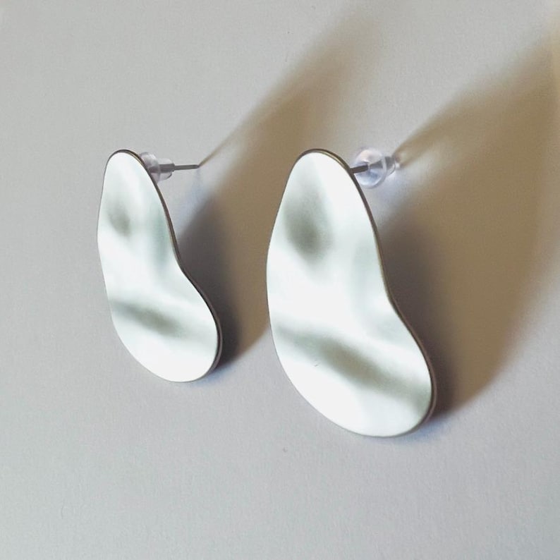 May include: A pair of silver-coloured, abstract-shaped earrings. Each earring has a smooth, reflective surface with a dark border. The earrings are attached to clear plastic posts.