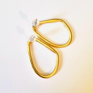 May include: A pair of gold-coloured, geometric earrings. Each earring has a unique, irregular shape with a rounded bottom and a straight top. The earrings have a post-back closure with a clear, rounded backing.