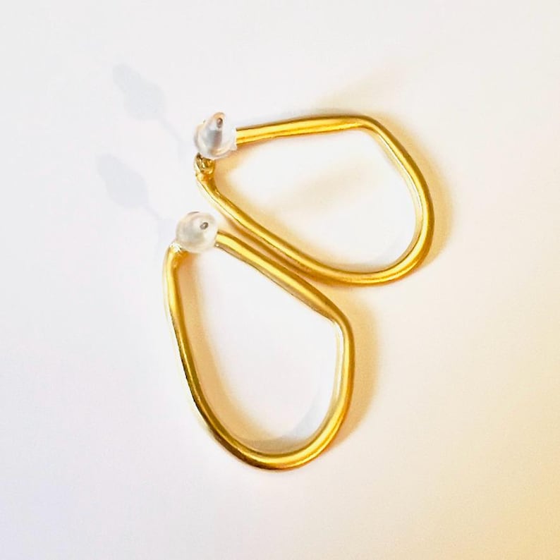 May include: A pair of gold-coloured, geometric earrings. Each earring has a unique, irregular shape with a rounded bottom and a straight top. The earrings have a post-back closure with a clear, rounded backing.