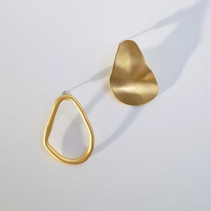 May include: A pair of gold-coloured earrings. One earring is a solid, irregular shape, while the other is an open, teardrop-like design. The earrings are set against a white background, with a shadow cast to the side.