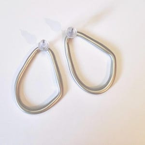 May include: A pair of silver-coloured, teardrop-shaped earrings. Each earring has a clear, rounded post at the top. The earrings are displayed on a white surface, with soft shadows.