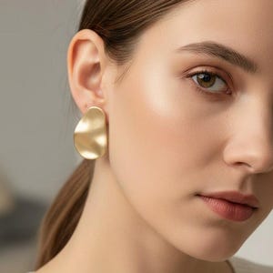 May include: Gold-coloured, abstract-shaped earrings. The earrings have a smooth, matte finish and a slightly irregular, organic form. The earrings are worn on the ear of a person.