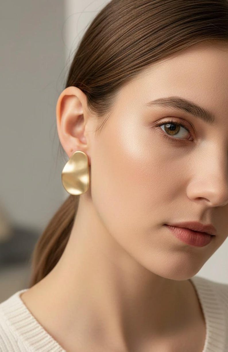 May include: Gold-coloured, abstract-shaped earrings. The earrings have a smooth, matte finish and a slightly irregular, organic form. The earrings are worn on the ear of a person.