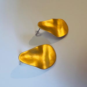 May include: A pair of gold-coloured stud earrings. Each earring has a unique, organic shape with a textured surface. The earrings are set against a light background, highlighting their metallic sheen and design.